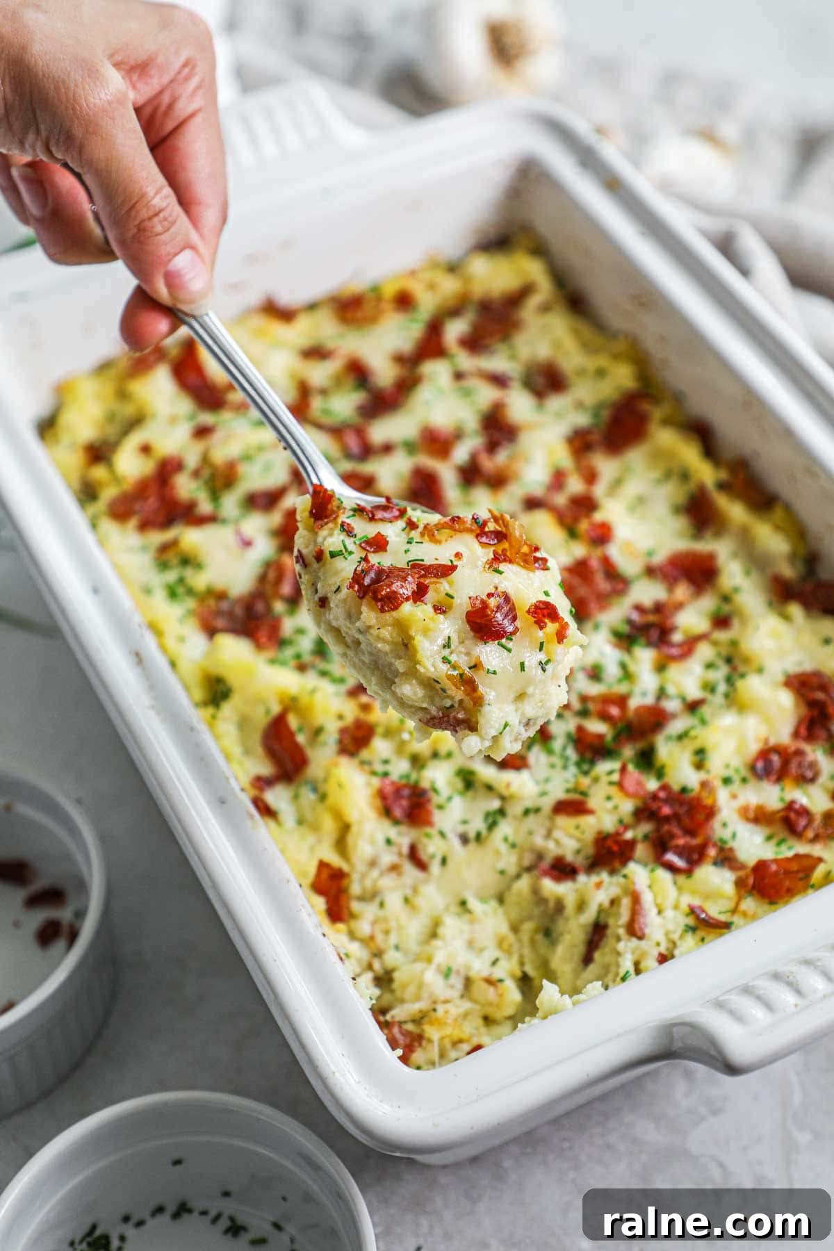 Creamy twice-cooked mashed potatoes with roasted garlic, gruyere, chives, and crispy prosciutto in a Le Creuset baker with a spoon holding a cheesy scoop.