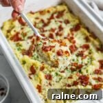 Creamy twice-cooked mashed potatoes with roasted garlic, gruyere, chives, and crispy prosciutto in a Le Creuset baker with a spoon holding a cheesy scoop.
