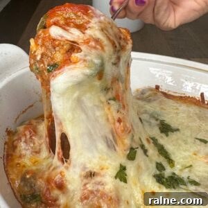 A hand with purple-painted nails lifts a cheesy, baked meatball from a casserole dish filled with melted cheese, tomato sauce, and garnished with chopped herbs.