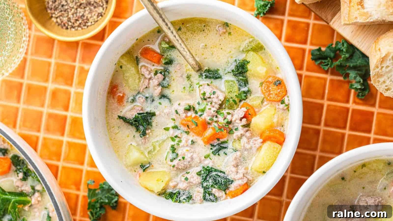 17 Comforting Soup Recipes for Quiet Evenings 18 A bowl of creamy soup with sausage, kale, carrots, and potatoes, with a spoon inside, on an orange tiled surface.