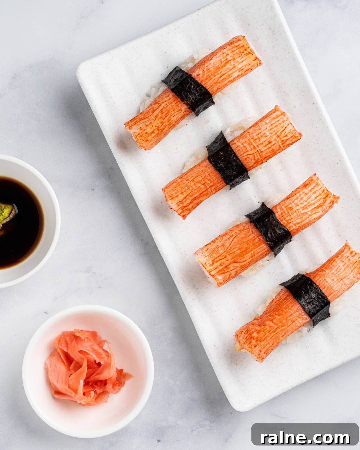 4 Kanikama nigiri sushi on a white plate with a side of soy sauce, wasabi and pickled ginger.