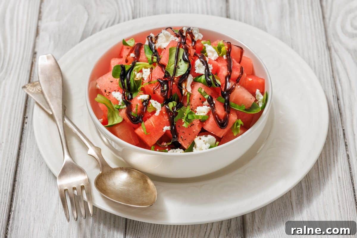 23 Make-Ahead Salads That Stay Fresh in the Fridge and Lunch Bag 7 A bowl of watermelon salad with feta cheese, arugula, and balsamic glaze, placed on a white plate with a fork and spoon on the side.