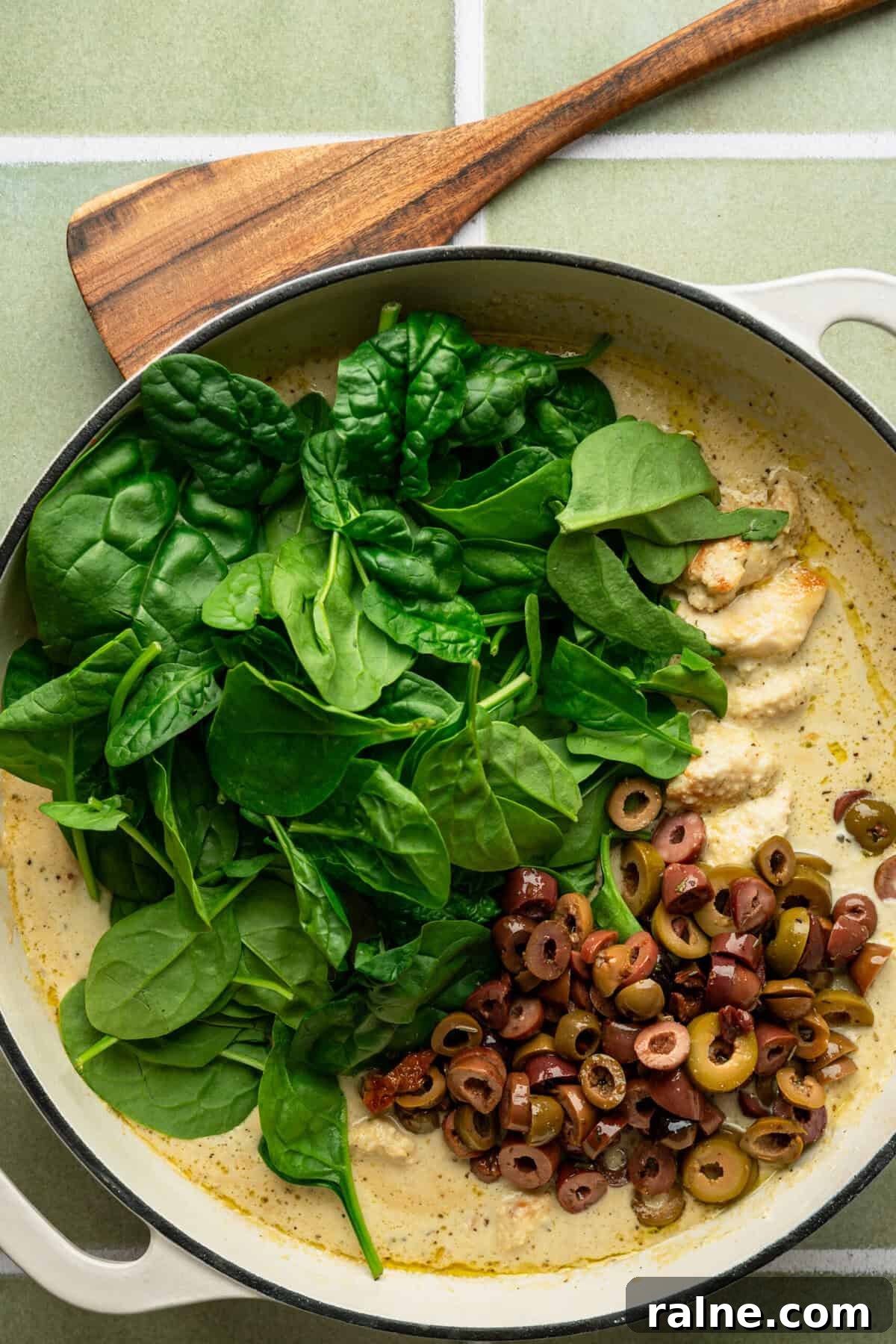 A pan filled with a white sauce and topped with fresh spinach and chopped black olives.