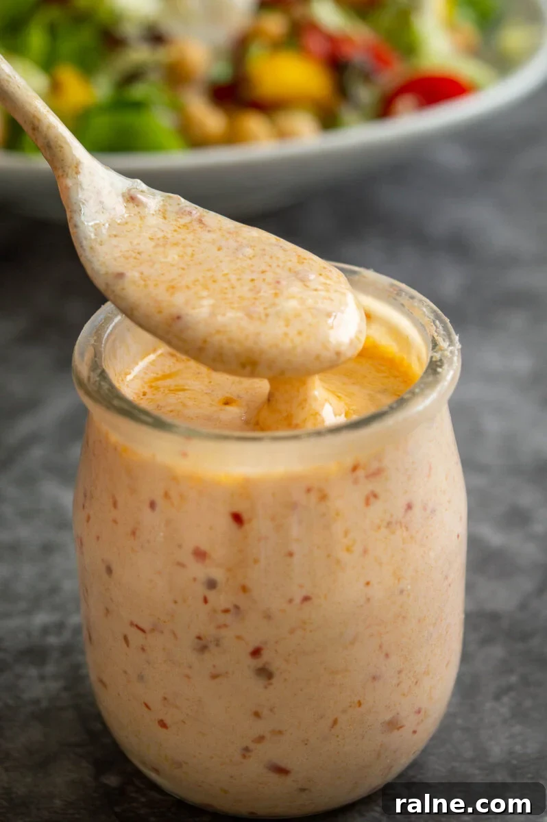 A silver spoon coated in creamy orange dressing over a small glass jar of it on a grey background.