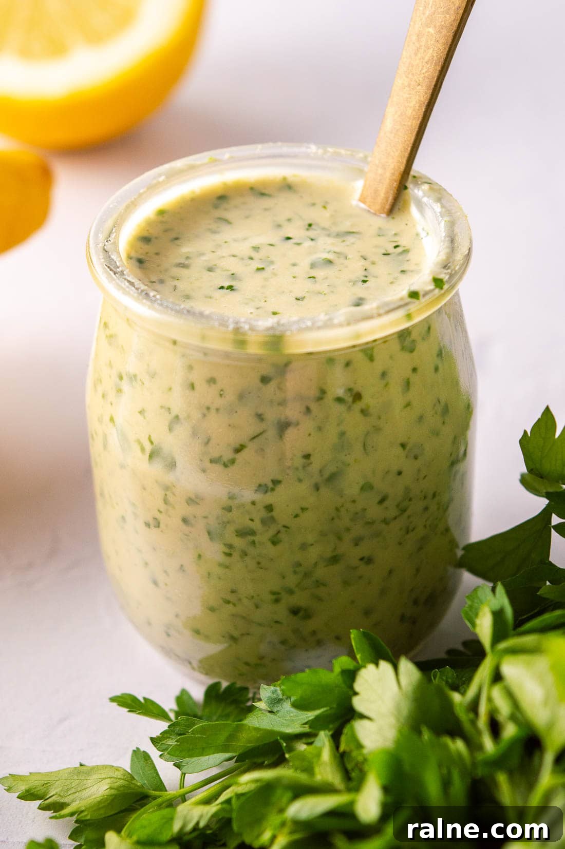 A glass jar with a pale herby sauce, lemons behind it and cilantro leaves in front of it.