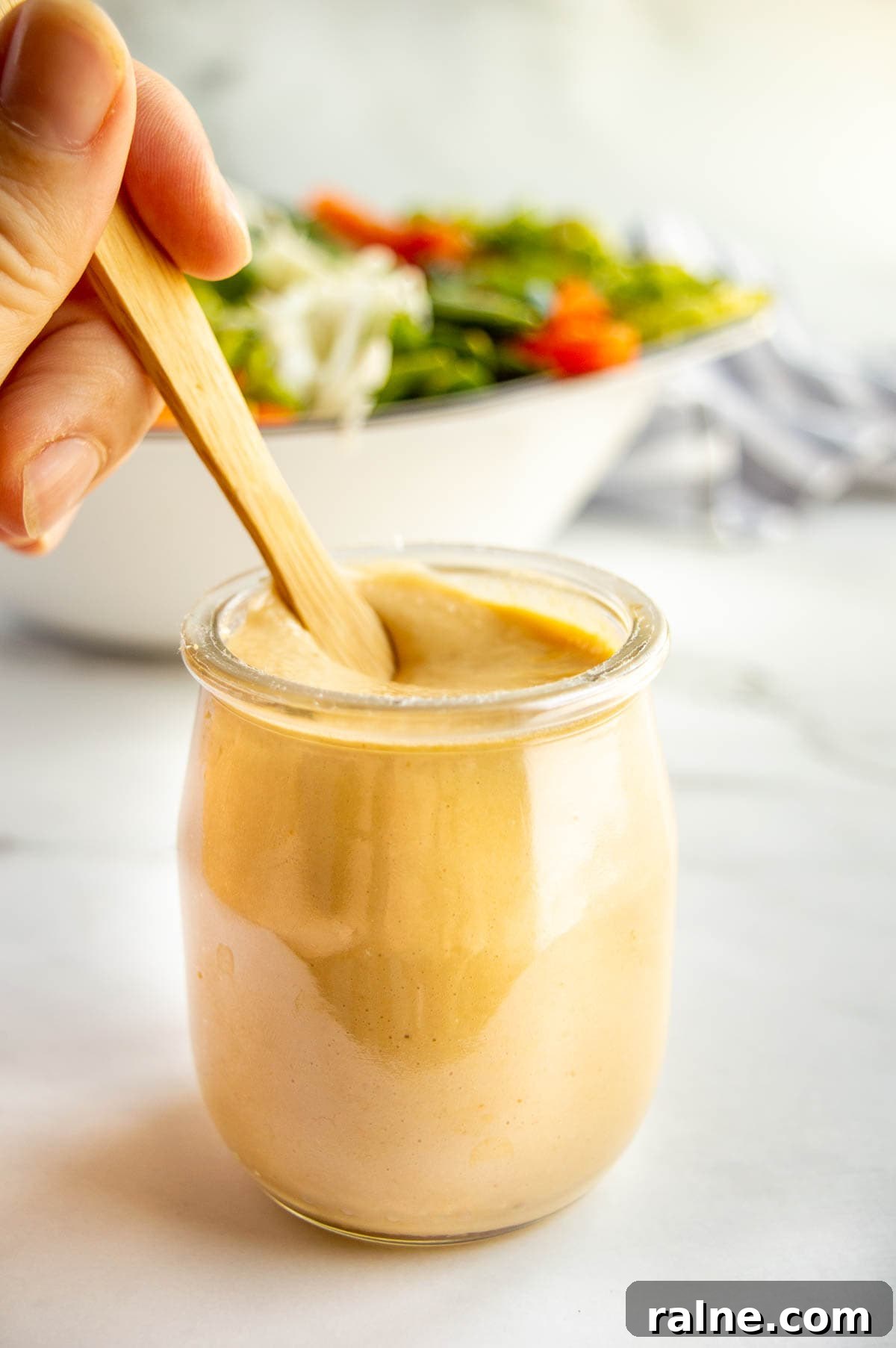 A glass jar with thick light brown dressing in it being stirred by a hand