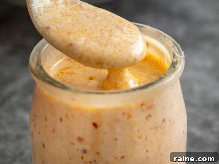 A silver spoon coated in creamy orange dressing over a small glass jar of it on a grey background.